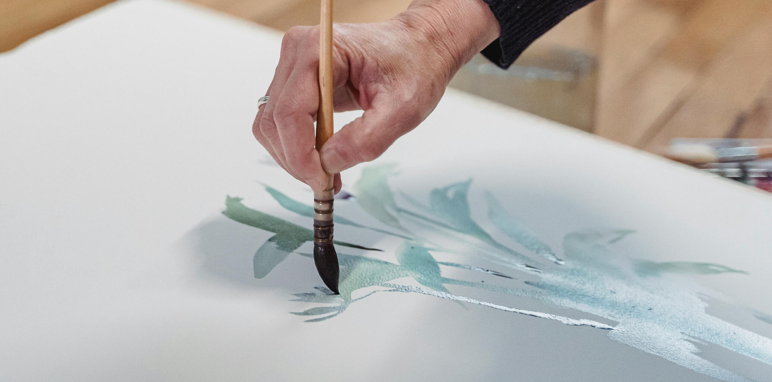 Person painting green leaves with watercolor on a white canvas.
