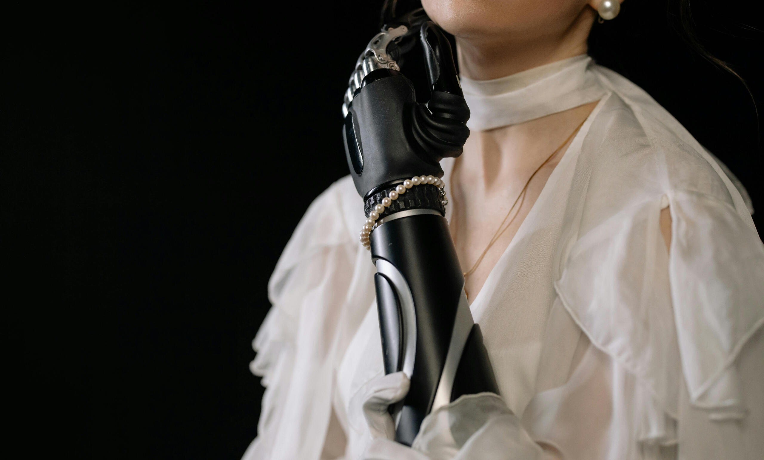 Woman wearing a white gown with a black prosthetic left arm, accessorized with pearl earrings and a pearl bracelet.
